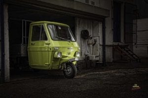 A green three-wheeled vehicle is parked in a dimly lit garage or industrial area. The vehicle, resembling a small truck, has a single wheel at the front and two at the back. Stairs and machinery are visible in the background, giving it a vintage feel perfect for a retro scooter shoot.