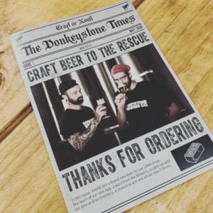 A vintage-style newspaper, "The Donkeystone Times," featuring a header with “Craft Beer to the Rescue” and an image by renowned Oldham photographer of two men toasting beer glasses. Below, the text reads, "Thanks for Ordering." Issue details and articles are also visible.