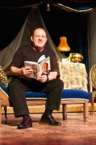 A man sitting on a blue couch holds an open book, seemingly reading it aloud. The background features a table with a floral tablecloth, a decorative lamp, various household items, and draped curtains. Capturing the scene's warm lighting and cozy ambiance is reminiscent of an Oldham photographer's touch.