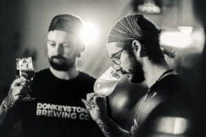 Two men in a dimly lit room, examining and tasting beer from glasses. Both have facial hair and wear dark t-shirts with "DONKEYSTONE BREWING CO" printed on them. The background features gentle lighting and blurred elements, adding to the ambiance, captured artfully by an Oldham photographer.