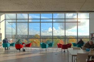 A spacious room with floor-to-ceiling windows showcasing a scenic view of a cloudy blue sky and autumn trees. Inside, people sit on colorful chairs by the windows, reading and working. The room, capturing an aesthetic that even an Oldham photographer would appreciate, features bright natural light and a modern design.