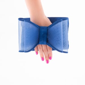A hand with pink nail polish holds a blue clutch bag designed in the shape of a large bow. Captured expertly by an Oldham photographer, the white background accentuates the bold color of the bag and contrasts sharply with the bright nails.