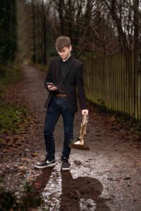 A young man dressed in a black blazer, black shirt, and jeans stands on a muddy path surrounded by trees. He looks at his phone in his right hand while holding a brass instrument, possibly a trumpet or French horn, in his left hand. Captured by a head shot photographer from Manchester, the path is lined with a wooden fence.