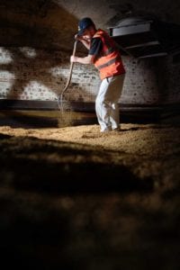 An Oldham photographer captures a worker in a high-visibility vest and cap, using a large shovel to turn and spread grains on the floor of a dimly lit, brick-walled industrial space. The scene, rich in shadows and light, emphasizes both the texture of the grains and the effort put forth by the worker.