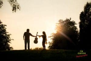 Silhouetted against a sunset, a man and a woman stand on a grassy hill. The man holds out a guitar to the woman. Trees frame the serene scene, while "Tim Simpson Photography - Oldham Photographer" is written in colorful text at the bottom right corner.