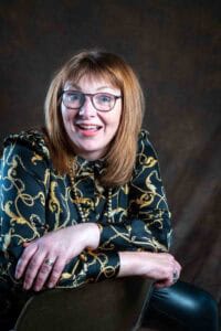 A woman with shoulder-length, light brown hair and glasses is facing the camera, smiling broadly. She is leaning forward on the back of a chair, wearing a patterned shirt with gold chains on a dark background. The backdrop, reminiscent of Manchester's vibrant music and comedy scene, is a dark, textured fabric.