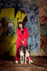 A woman stands in front of a colorful graffiti wall in Manchester, wearing a bright red suit. She is holding leashes for two black and white dogs standing beside her. The scene is illuminated with vibrant light, highlighting the intricate graffiti art and the woman's confident pose, almost as if captured in a moment of music and comedy photography.