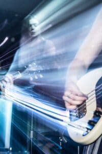 A dynamic image of a music performance in Manchester showing a blurry, ethereal motion effect. A guitarist in the background and a bassist in the foreground are seen playing their instruments with colorful light streaks creating a sense of energetic movement, captured through music and comedy photography.