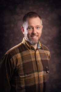 A man with short hair and a beard smiles at the camera. He is wearing a brown plaid shirt with front pockets. The background, reminiscent of Manchester's moody ambiance, is dark with a soft, mottled texture. The lighting highlights the right side of his face and adds depth to the image.
