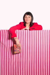 A woman wearing a bright red blazer stands behind a pink and white striped surface, resting her arms on it. She holds a colorful, patterned book in her right hand, with a neutral expression on her face. The background is plain white, reminiscent of minimalist Manchester photography styles.
