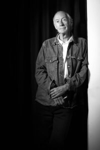 Black and white photograph of an older person standing against a dark backdrop. They are wearing a denim jacket over a button-down shirt, holding a cigarette. With a calm, contemplative expression, the dramatic contrast in light and shadow embodies the unique atmosphere of Manchester's music and comedy scenes.