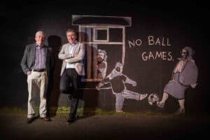 Paul Heathcote and his father stand in front of a wall mural in Manchester featuring an old lady leaning out of a window with the words "No Ball Games" painted beside her. Children are depicted playing with a ball below. Both men, smiling and dressed in suits, could easily be stars of music or comedy. The scene is lit by a spotlight.
