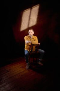 A person with a short beard is sitting on a wooden chair in a dimly lit room with a single window casting light on the wall behind. The person, perhaps taking a break from music and comedy photography in Manchester, is wearing a mustard-colored jacket, blue jeans, and holding folded arms while leaning forward.