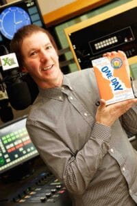 Clint Boon wearing a checkered shirt is standing in a recording studio in Manchester, holding up a book titled "One Day" by David Nicholls. They are smiling, surrounded by music and recording equipment, including a microphone and control board.