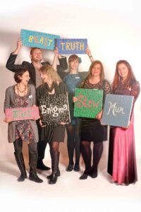 A group of six people stand together holding colorful signs with words on them. The signs read: "BEAST," "TRUTH," "Enigma," "Slaw," "Eat," and "Mix." The group is smiling and appears to be enjoying the moment, as a photographer captures this lively scene in Manchester, blending elements of music and comedy.
