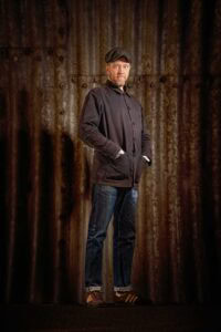 A man stands against a rusted corrugated metal background. He is wearing a flat cap, jacket with pockets, jeans, and sneakers. His hands are in his jacket pockets, and he has a calm expression on his face. The lighting is warm and focuses on him, capturing the essence of Manchester's music scene.