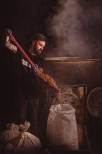 A person with tattoos and a beard, wearing a black shirt and gloves, uses a red shovel to pour grains from a large industrial container into a white bag. Thick steam or smoke fills the dimly lit room, creating an atmosphere that any Oldham photographer would find rustic and industrial.