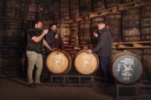 Three men are standing in a dimly lit room with wooden barrels on racks. Each man is holding a glass of dark liquid, presumably whiskey, and appears to be tasting it. The barrels in front have labels indicating different years and types of malt whiskey from Spirit of Yorkshire, resembling a brewery gallery.