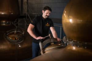 A person in a black shirt works at a distillery, inspecting a large copper still. The background resembles a brewery gallery, featuring more equipment and industrial piping. The mustached individual is focused on the task at hand in the dimly lit environment.