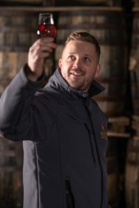 A person wearing a gray jacket inspects a glass of red liquid, holding it up towards the light. They are standing in front of stacked wooden barrels in the brewery gallery, looking up at the glass with a focused expression.
