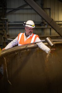 A man wearing a white construction helmet, safety glasses, and an orange safety vest stands inside an industrial facility. He is observing and touching a large metal tank in what appears to be a brewery while water or liquid flows from a spout. The background shows various industrial equipment.
