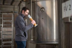 A man in a gray jacket examines a tall cylinder filled with a light brown liquid, standing next to a large stainless steel tank in the brewery gallery. Metal shelves and another tank labeled "FV 2" are visible in the background.