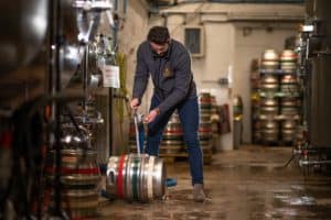 A man wearing a gray jacket and jeans is operating a keg in a brewery gallery filled with various barrels and brewing equipment. He is connecting a hose to the keg while standing on a wet industrial floor. The background features stacks of colorful kegs and pipes.