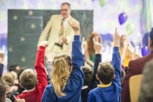 Children in a classroom sit on the floor with their hands raised, looking toward an adult standing at the front. The adult appears to be leading a lesson or activity. Balloons and decorative elements can be seen in the background, suggesting a festive atmosphere, captured perfectly by an Oldham photographer.
