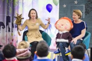 Two women are seated in front of a group of children. One woman is holding a brown owl puppet and gesturing animatedly, while the other woman is holding a large doll with orange hair. The colorful setting, captured beautifully by an Oldham photographer, features balloons and animal illustrations.