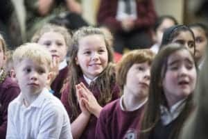 A group of young children, both boys and girls, sit closely together, appearing attentive and engaged. Captured by an Oldham photographer, they wear school uniforms—some in maroon sweaters and others in white shirts. One girl near the center is smiling brightly with hands clasped.
