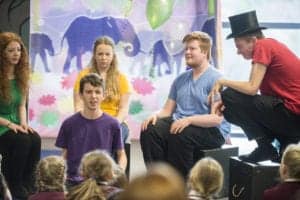 A group of young people are performing a skit in a brightly decorated room. Captured by an Oldham photographer, one person in a purple shirt is speaking, with others seated around him looking attentive. One performer wearing a top hat sits on a box. The background features colorful elephant silhouettes.