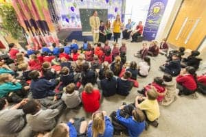 A group of children sit on the floor facing a stage where several adults are standing and talking. The room, captured beautifully by an Oldham photographer, is decorated with colorful curtains and banners. The children are dressed in various uniforms, suggesting a school or community event.