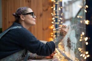 A person with tattoos on their arms, wearing glasses and a sweater, paints a design on a window decorated with string lights. They appear focused, with their hand raised, holding a paintbrush, and leaning close to the window; all while music fills the background, adding an artistic essence to this Manchester scene.