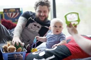 A bearded man with tattoos lies on a cushion, smiling at a baby who is sitting next to him holding toys. Another person is holding up a toy in front of the baby. A basket of stuffed animals and colorful toys is also present. Captured by an Oldham photographer, they are indoors, near a window.