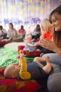 A group of adults and babies attend a sensory play session captured beautifully by an Oldham photographer. A baby lying on a pillow holds a colorful toy while an adult, sitting nearby, claps and smiles. The room is decorated with vibrant colors, and other participants are engaged in playful activities in the background.