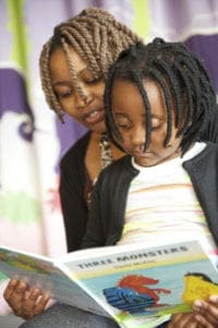 An adult with braided hair reads a brightly illustrated book titled "Three Monsters" with a child who has beaded cornrows. Captured by an Oldham photographer, the scene is warm and intimate, with a softly colored background suggesting a cozy environment suitable for reading.