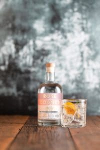 A bottle of Rose County Gin with a wooden cap sits on a wooden surface against a textured dark background, resembling an exhibit in a brewery gallery. Beside it is a clear glass filled with a drink, ice, and an orange slice garnish.