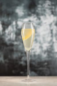 A champagne flute filled with a light-colored beverage and garnished with a lemon twist, set against a dark, blurred background, showcases the elegance of this brewery creation.