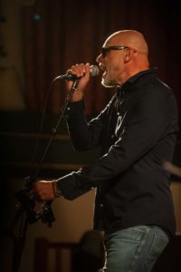 A man with a shaved head and sunglasses sings passionately into a microphone while gripping a stand. He is wearing a dark long-sleeved shirt and jeans, captured brilliantly by the UK's best event photographer under warm stage lighting against a dark background.