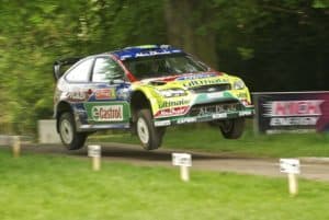 A rally car with colorful branding, including Castrol and Abu Dhabi, is mid-air during a jump on a forested dirt track. Captured by the UK's best event photographer, numbered track markers on green grass flank the car, and blurred banners can be seen in the background.