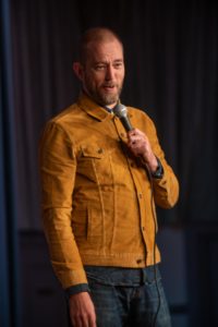 A bald man with a beard, wearing a mustard-colored jacket and holding a microphone, stands on a stage. The background is dimly lit with curtains, suggesting a performance or comedy show setting. Captured by the UK's best event photographer, the scene exudes anticipation and charm.