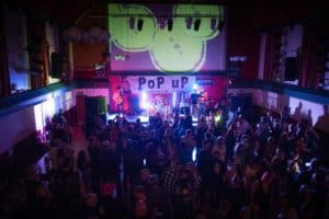A lively crowd watches a band perform on stage in a dimly lit venue. The band is under a "PoP uP" banner, and the backdrop features green illuminated designs. Captured by the UK's best event photographer, people are standing and dancing in front of the stage, enjoying the performance.