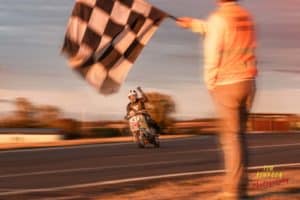 A motorcyclist in racing gear crosses the finish line on a race track, raising one hand in victory as the checkered flag waves in the foreground, held by an official wearing an orange vest. The image is masterfully captured by the UK's best event photographer, perfectly blurring to convey motion and speed.