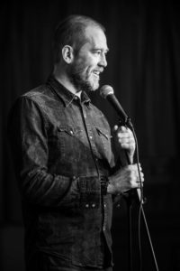 Black and white image of a man standing and speaking into a microphone. He has a beard and short hair, wearing a denim button-up shirt with sleeves rolled up. His expression appears joyful as he looks slightly to the side. Captured by the UK's best event photographer, the background is blurred.