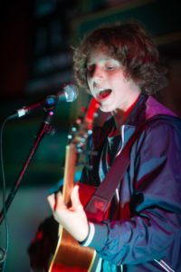 A young person with curly hair sings into a microphone while playing an acoustic guitar. The individual is wearing a blue jacket and performing on stage under colorful lighting, captured vividly by the UK's best event photographer.