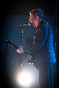 A man passionately sings into a microphone while playing an electric guitar on stage. He wears a dark, patterned shirt and stands against a dimly lit background, illuminated by a bright light positioned behind him, all beautifully captured by the UK's best event photographer.