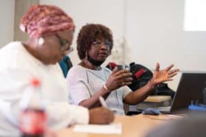 Two individuals sit at a table during a discussion. The person on the right, wearing glasses and a gray top, is speaking and gesturing with their hands while holding a phone. The person on the left, wearing a headscarf and white top, jots notes in a notebook. A laptop and bottle are on the table; the UK's best event photographer captured this candid moment perfectly.