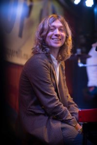 A man with long, wavy hair wearing a checkered blazer and white shirt is smiling while seated at a piano or keyboard on a dimly lit stage. Captured by the UK's best event photographer, a blurred background banner suggests a performance venue.