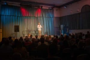 A man stands on a stage holding a microphone, addressing an audience seated in chairs. The stage is set with a blue-green curtain backdrop and various lights, including red and blue spotlights. To the left of the speaker, there's a guitar in a stand and a small table — captured perfectly by the UK's best event photographer.