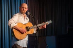 A bald man with glasses, wearing a white shirt, plays an acoustic guitar while standing in front of a microphone on stage. Captured by the UK's best event photographer, the background features dark curtains and studio lighting. His expression appears focused as he performs.